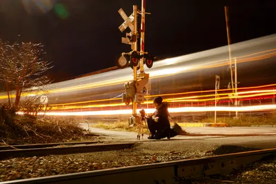 At night, a person kneels on a gravel roadside next to a railroad crossing signal, looking through a camera mounted on a tripod. Behind them, a passing train is rendered as a blur of long, horizontal light trails against the dark background.