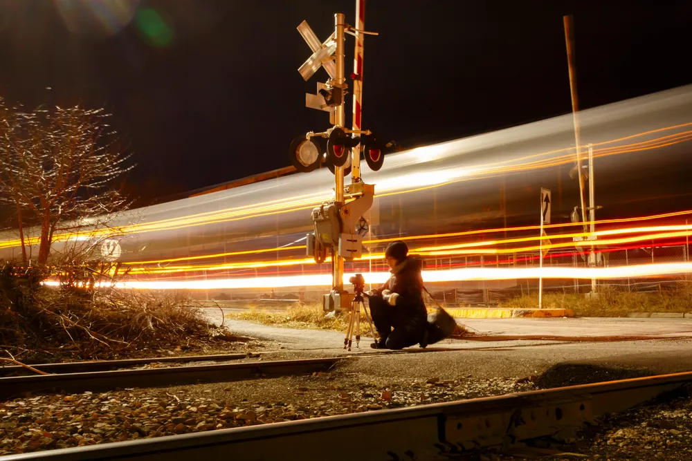 At night, a person kneels on a gravel roadside next to a railroad crossing signal, looking through a camera mounted on a tripod. Behind them, a passing train is rendered as a blur of long, horizontal light trails against the dark background.