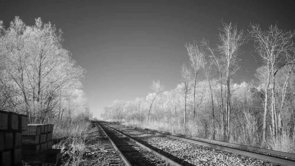 An infrared black and white photograph of railroad tracks extending toward the horizon. The tracks are flanked by trees and bushes with white, ethereal-looking foliage, under a stark, dark sky. A stack of wooden ties sits on the left side of the frame.